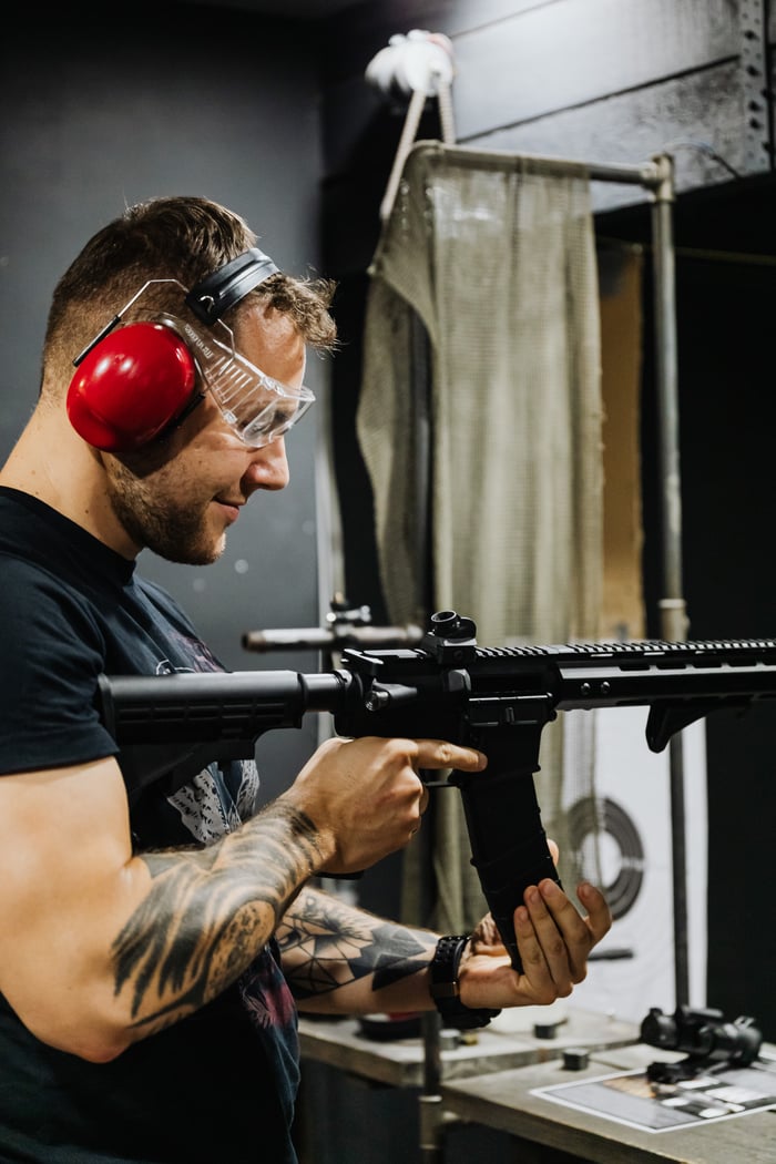 Tattooed man reloading rifle at indoor shooting range, wearing earmuffs and goggles.